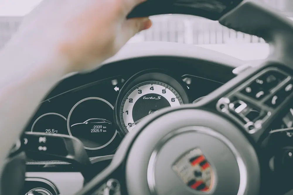Mão do condutor no volante, com os mostradores e o emblema Porsche no cubo