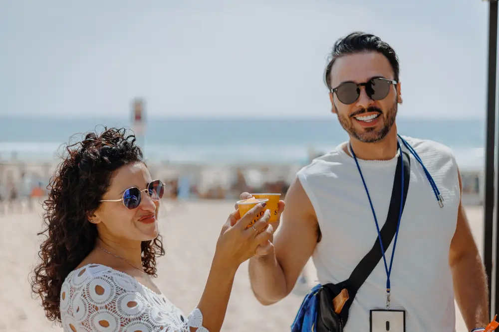 Dois festivaleiros com bebida na mão, com a praia ao fundo