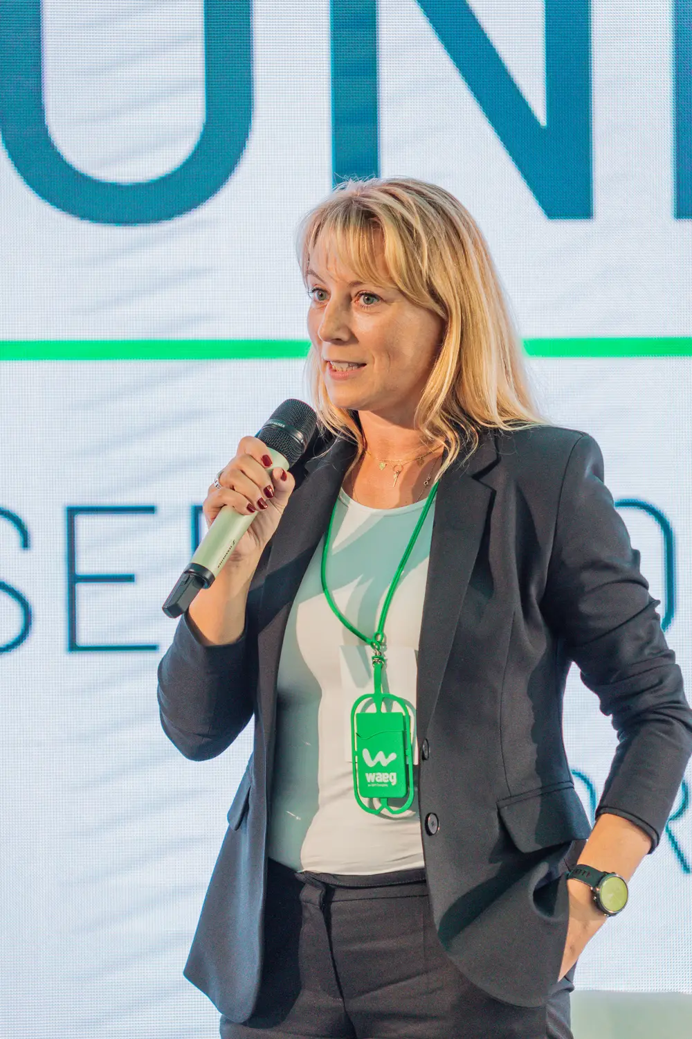Oradora de blazer com microfone, diante do cenário da conferência